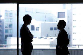 Besprechung zweier dunkler Silhouetten vor einer Glasfront mit Blick auf Bürogebäude im Hintergrund