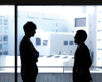 Besprechung zweier dunkler Silhouetten vor einer Glasfront mit Blick auf Bürogebäude im Hintergrund