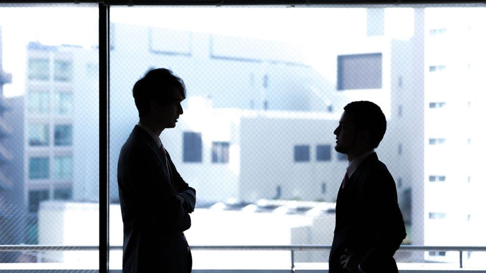 Besprechung zweier dunkler Silhouetten vor einer Glasfront mit Blick auf Bürogebäude im Hintergrund
